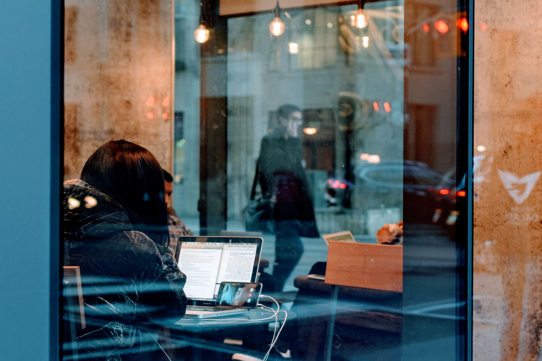 how to reset network settings in windows 10 - A person working on a laptop inside a café, viewed through a window with street reflections.