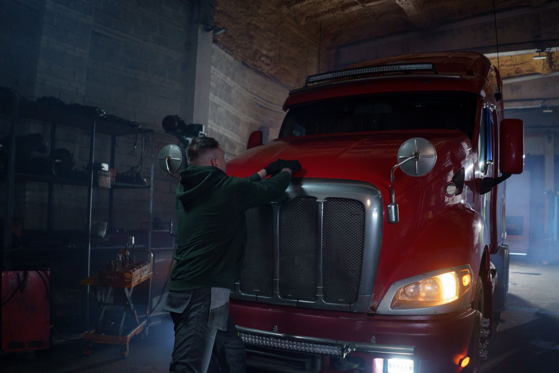 route 19 heavy truck repair - A mechanic in a dimly lit garage working on a red semi truck, focus on repair.