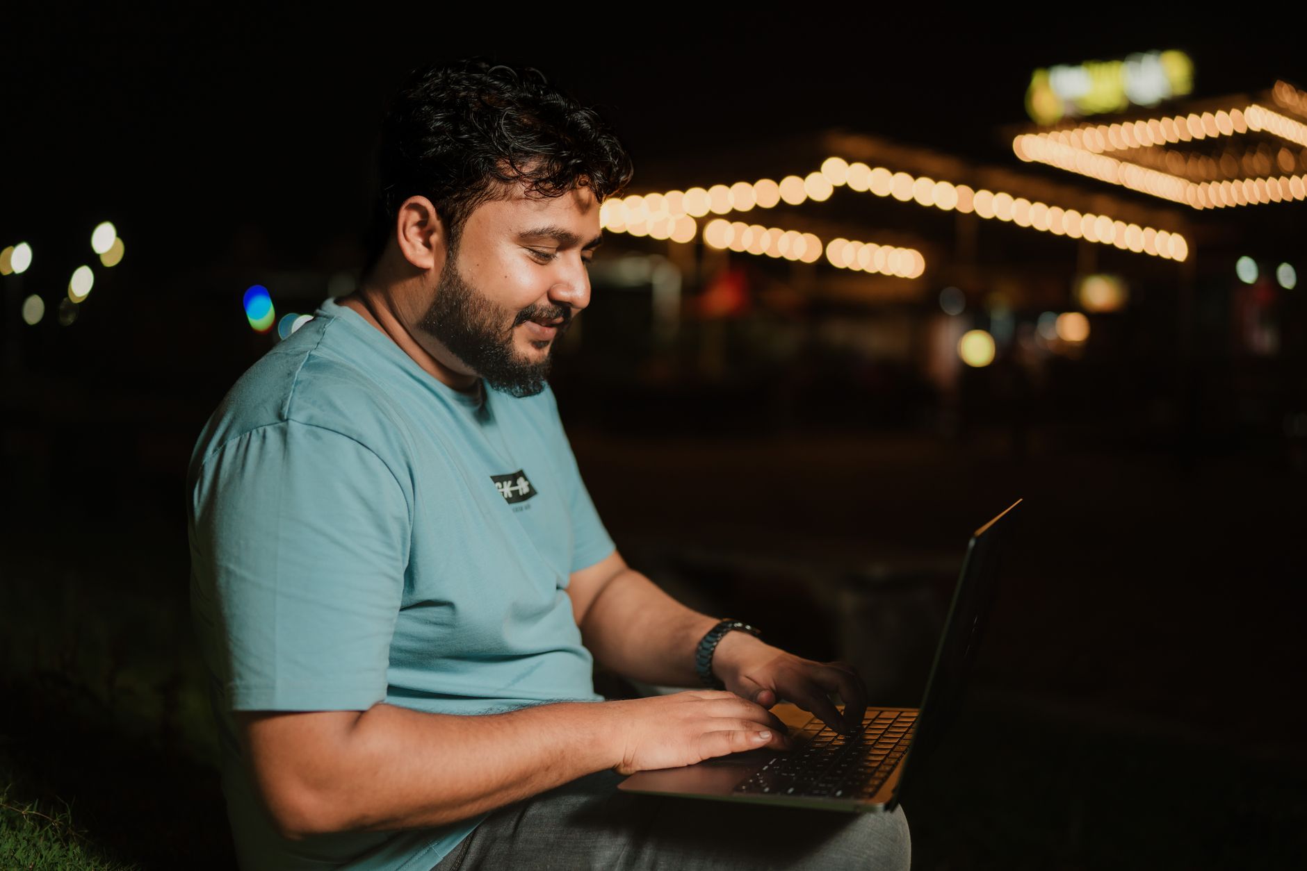 why won't my laptop connect to the wifi - A man in a blue t-shirt typing on a laptop outdoors under string lights at night.