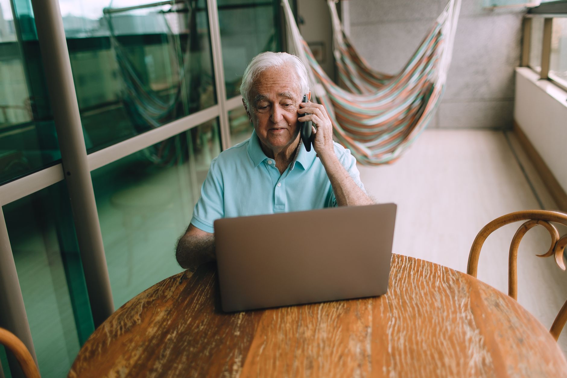 wifi is working on phone but not on laptop - Elderly man multitasking on laptop and phone, sitting at a wooden table indoors with hammocks in the background.